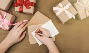 Ręka pisze kartkę, by stworzyć zaproszenie na urodziny. Wokół leżą prezenty owinięte w papier i ozdobione wstążkami.