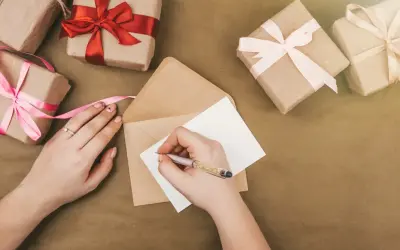 Ręka pisze kartkę, by stworzyć zaproszenie na urodziny. Wokół leżą prezenty owinięte w papier i ozdobione wstążkami.