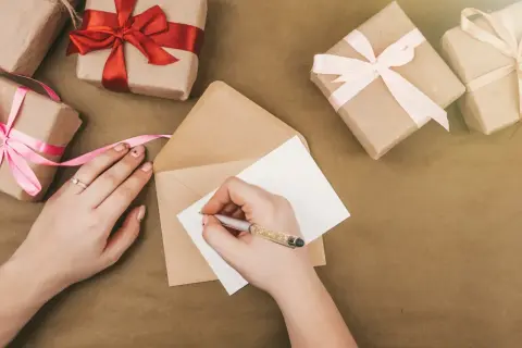 Ręka pisze kartkę, by stworzyć zaproszenie na urodziny. Wokół leżą prezenty owinięte w papier i ozdobione wstążkami.