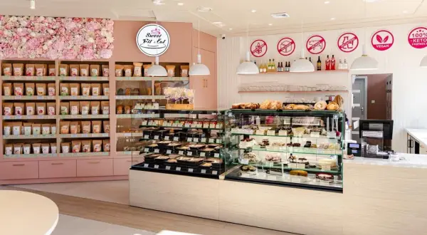 Wnętrze cukierni "Sweet Fit & Eat" z witrynami pełnymi ciast i ciasteczek. Reklama cukierni podkreśla opcje bez glutenu, cukru i laktozy.