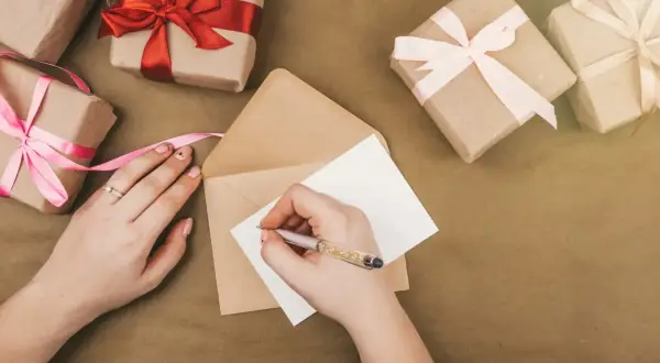 Ręka pisze kartkę, by stworzyć zaproszenie na urodziny. Wokół leżą prezenty owinięte w papier i ozdobione wstążkami.