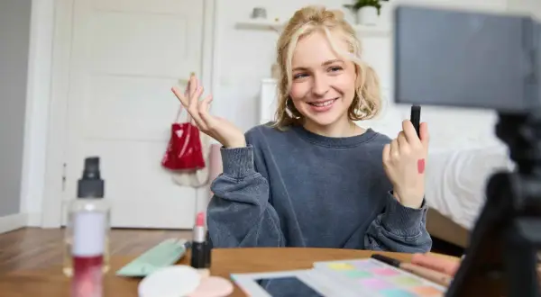 Uśmiechnięta influencerka z pomadką na dłoni pokazuje, jak zostać ambasadorem marki, nagrywając tutorial makijażowy.