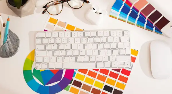 Na biurku grafika: klawiatura, myszka, okulary, słuchawki, koło barw i próbki kolorów, w tym **kolor pantone**.