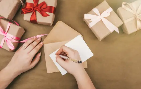 Ręka pisze kartkę, by stworzyć zaproszenie na urodziny. Wokół leżą prezenty owinięte w papier i ozdobione wstążkami.