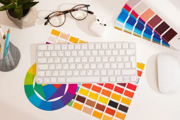 Na biurku grafika: klawiatura, myszka, okulary, słuchawki, koło barw i próbki kolorów, w tym **kolor pantone**.